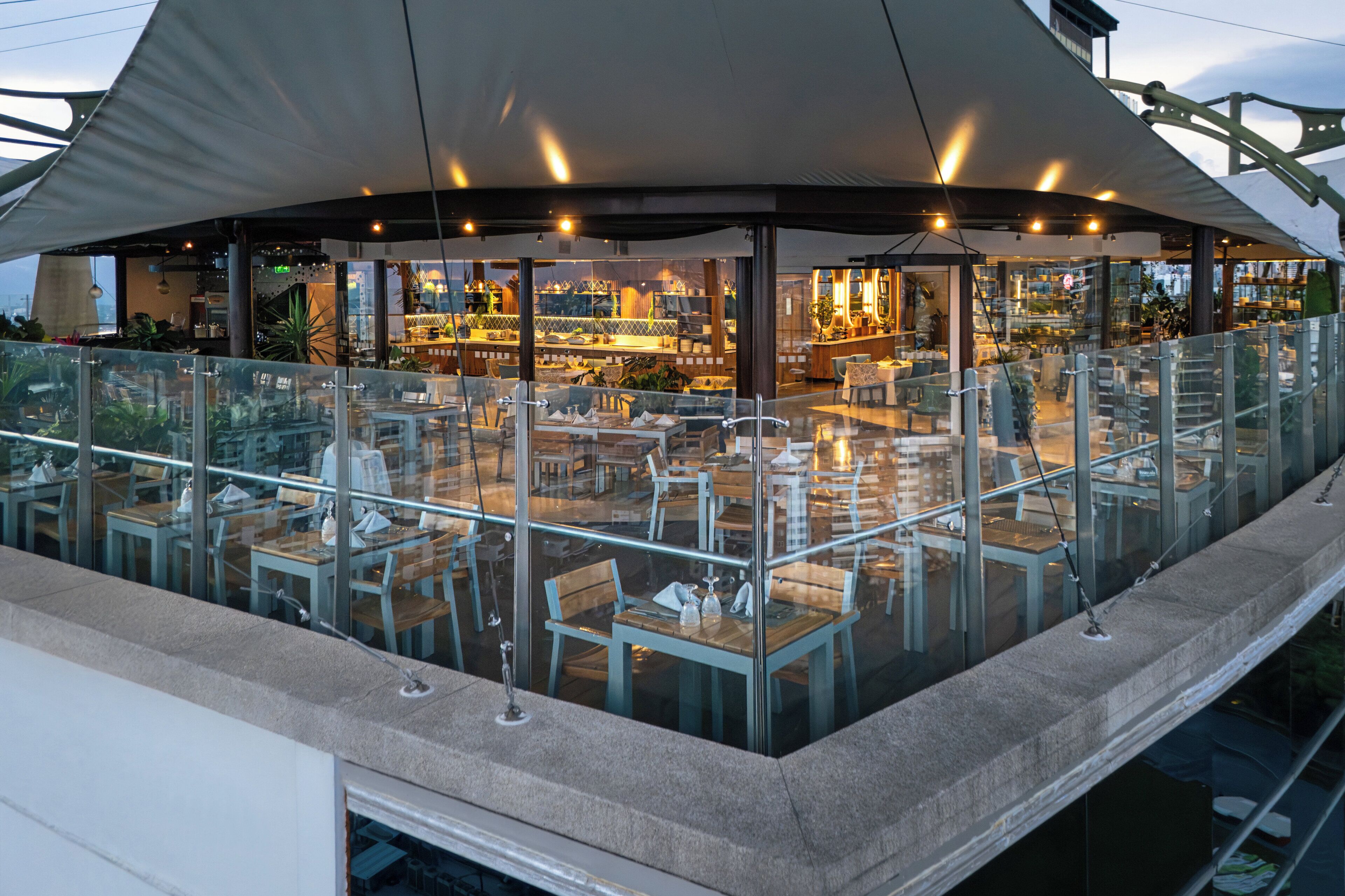 Un área de comedor al aire libre con barandillas de vidrio, mesas y sillas, y vista al espacio interior del restaurante.