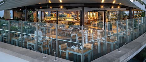 Un área de comedor al aire libre con barandillas de vidrio, mesas y sillas, y vista al espacio interior del restaurante.