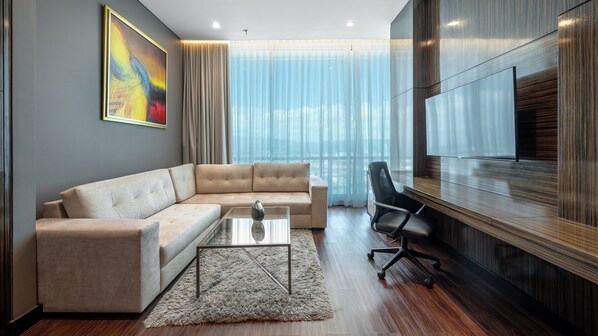 Ein modernes Wohnzimmer mit einem Ecksofa, einem Glastisch, einem Teppich, einem an der Wand montierten Fernseher und einem großen Fenster mit transparenten Vorhängen.