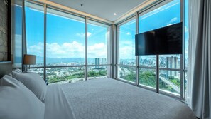 A hotel room with a large bed, a flat-screen TV, and a city view through floor-to-ceiling windows.