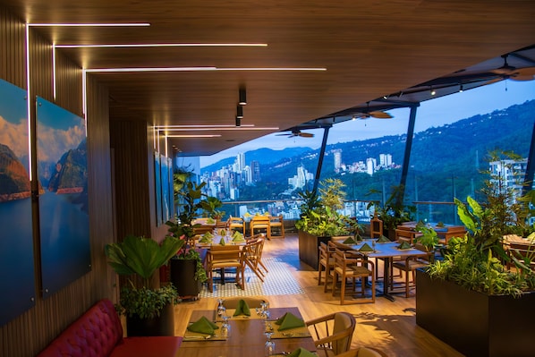 A rooftop restaurant with wooden furniture, potted plants, and a city view.