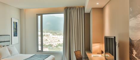 A hotel room with a bed, a desk with a chair, a television, and a large window with curtains.