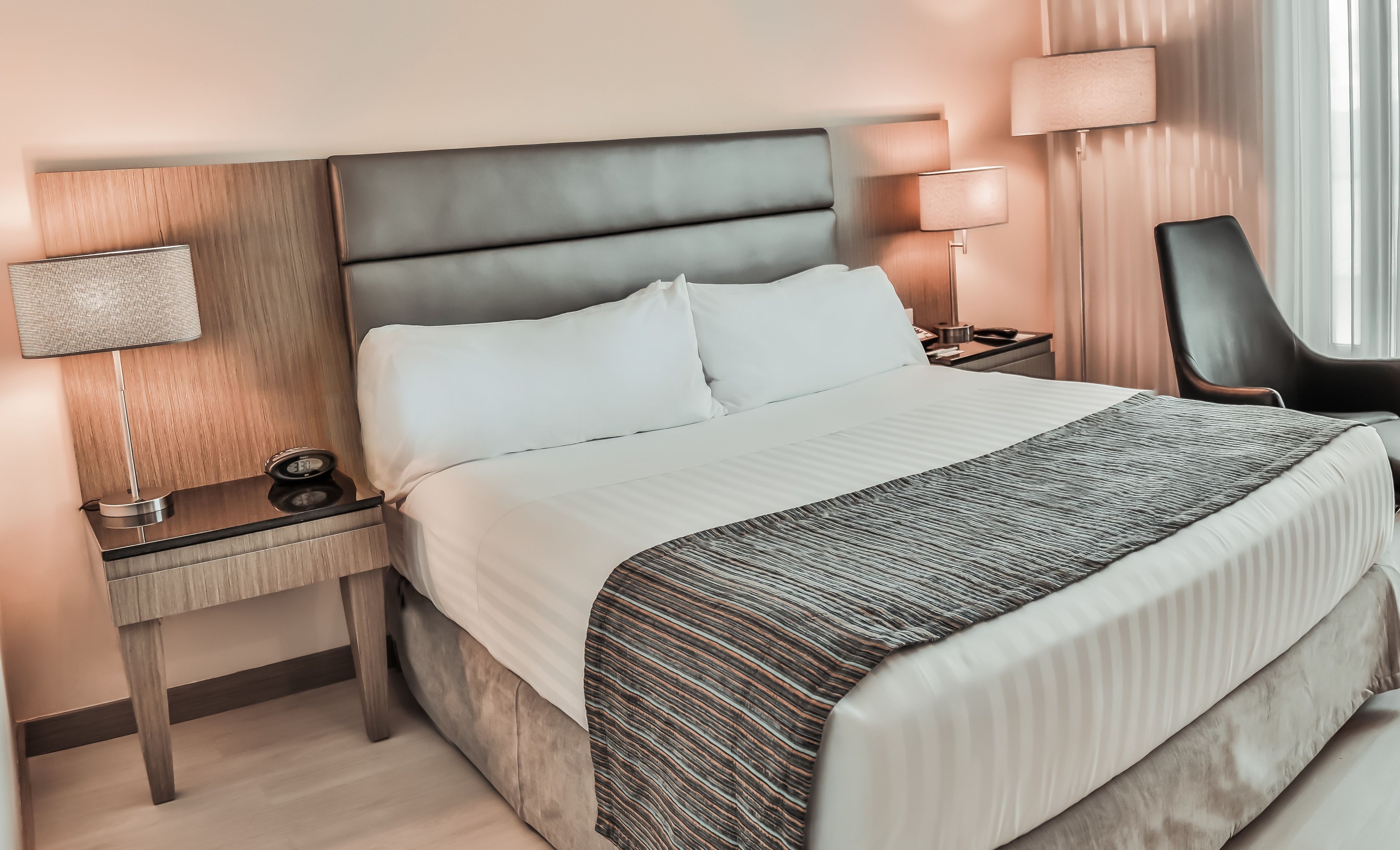 Ein ordentlich bezogenes Bett mit weißen Leinen, ein grau gepolsterter Kopfteil und eine gemusterte Decke. Ein Nachttisch mit Lampe und einer Uhr, und ein Stuhl in der Ecke.