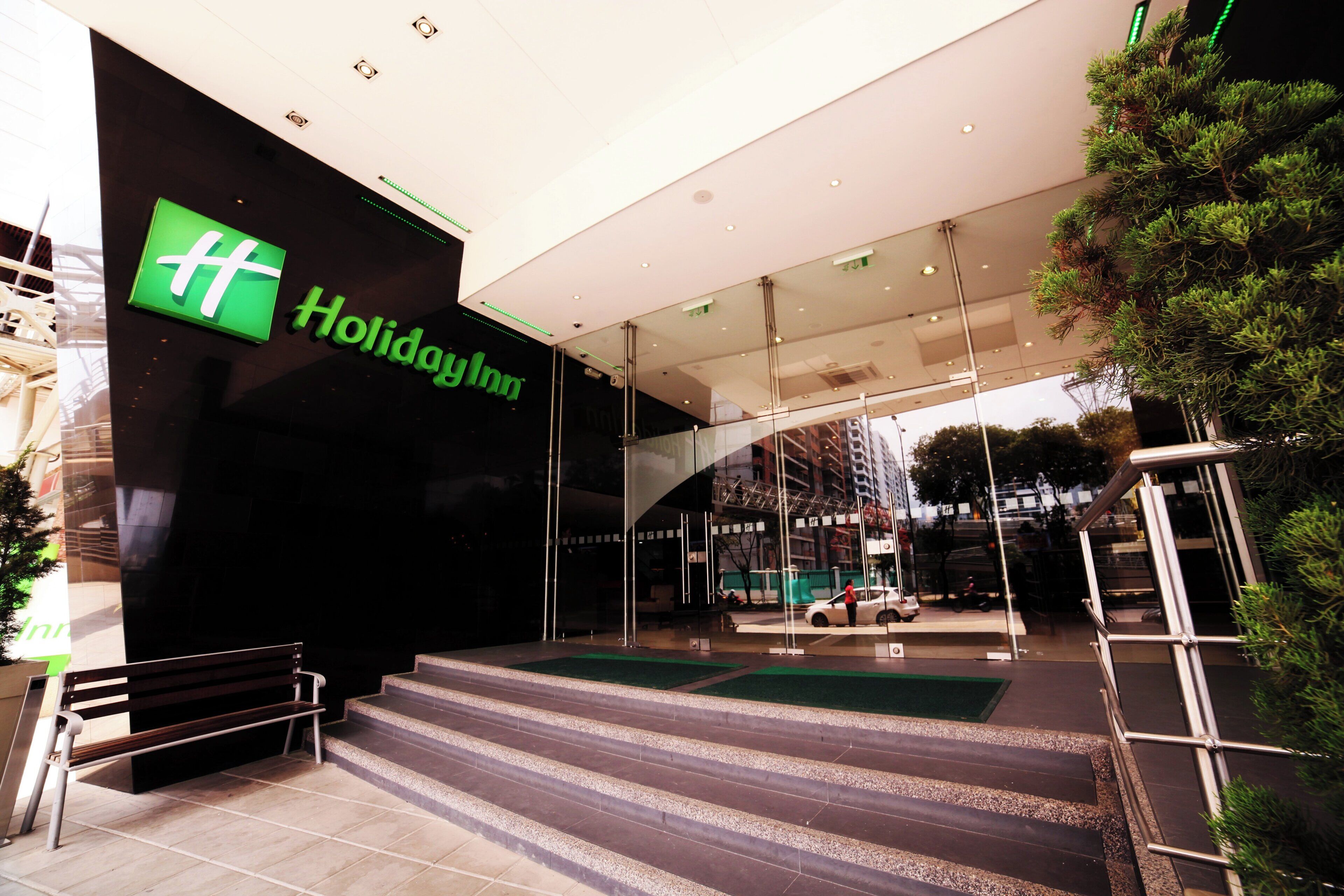 The exterior of a Holiday Inn hotel with a prominent green logo, a glass facade, and a staircase leading to the entrance.