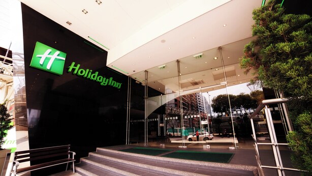 L’extérieur d’un hôtel Holiday Inn, avec son logo vert bien visible, une façade en verre et un escalier menant à l’entrée.