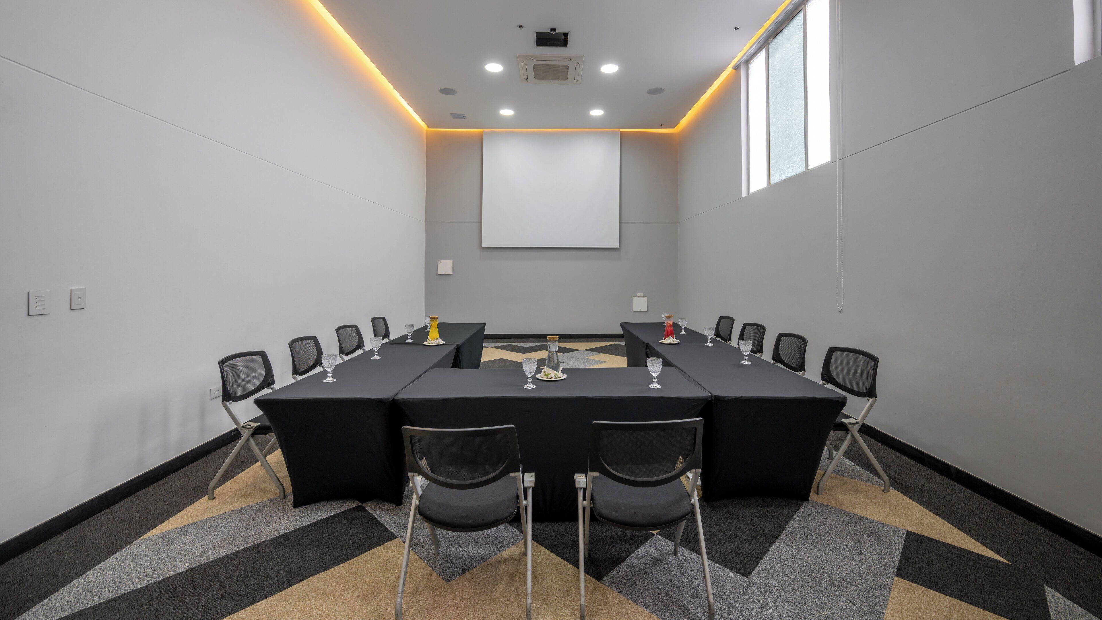 Una sala de conferencias con una mesa larga, sillas, una pantalla de proyector y piso alfombrado.
