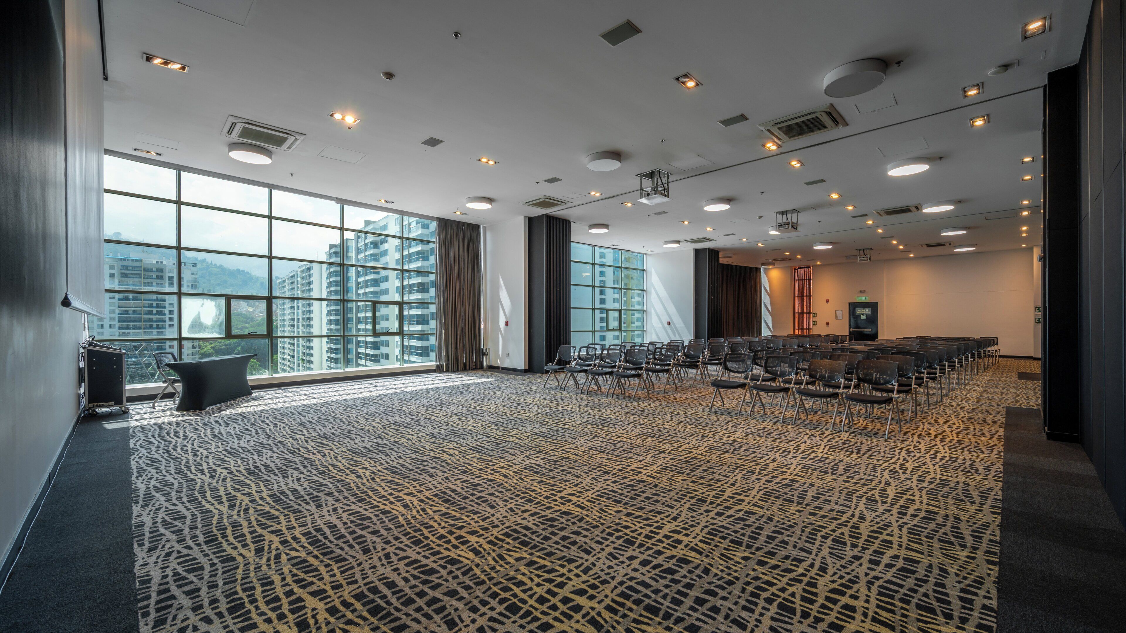 Una sala de conferencias con alfombra estampada, filas de sillas, una pantalla de proyector y grandes ventanales con vista a la ciudad.