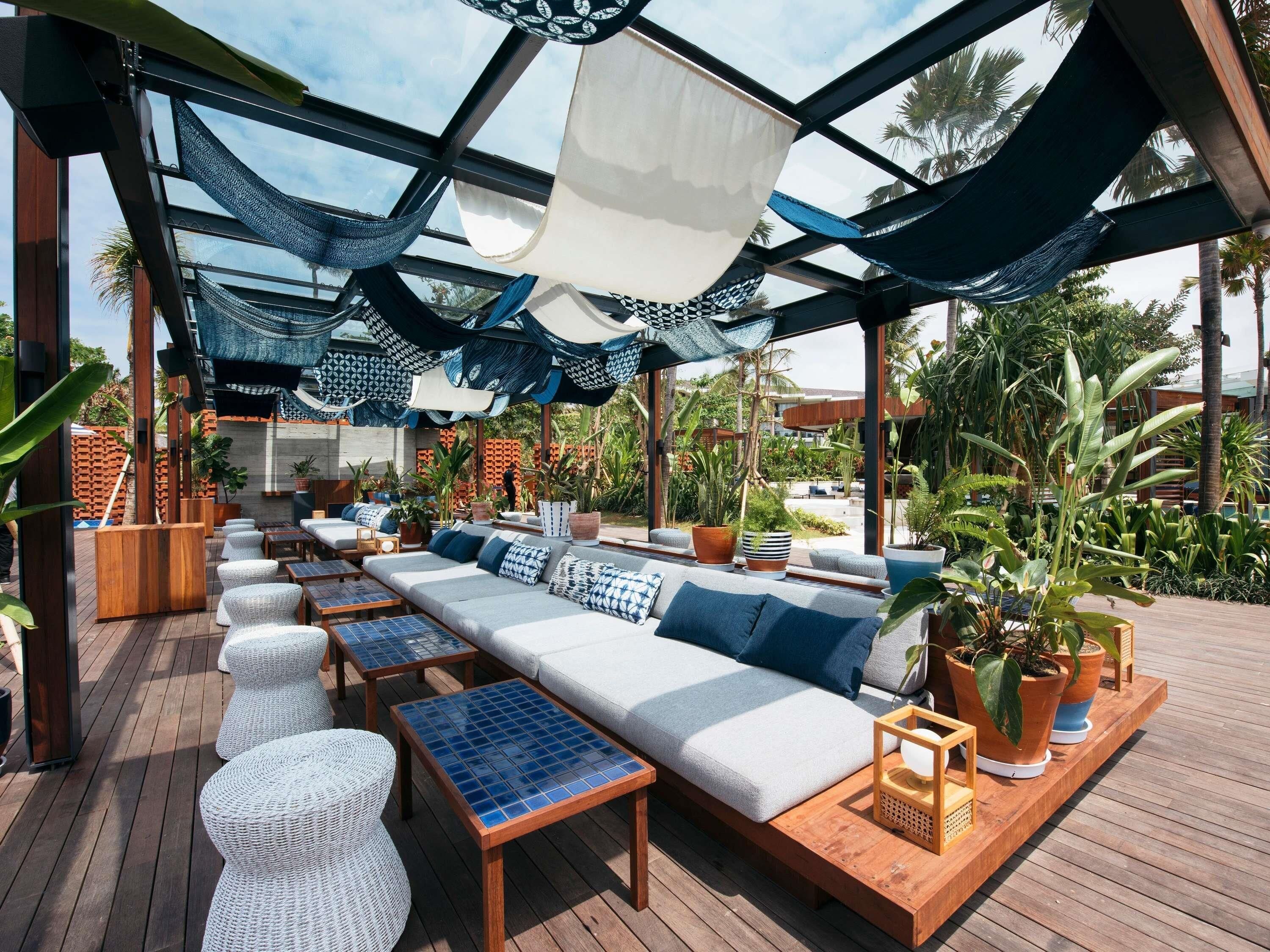 Un espace de détente extérieur comprenant un long canapé, des fauteuils confortables et des tables, le tout entouré de plantes en pot et agrémenté d’une terrasse en bois.