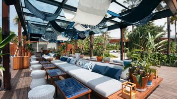 Un espace de détente extérieur comprenant un long canapé, des fauteuils confortables et des tables, le tout entouré de plantes en pot et agrémenté d’une terrasse en bois.