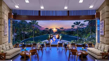 Ampla área de estar com vista para um pôr do sol sobre um jardim paisagístico, mobiliada com sofás e poltronas.