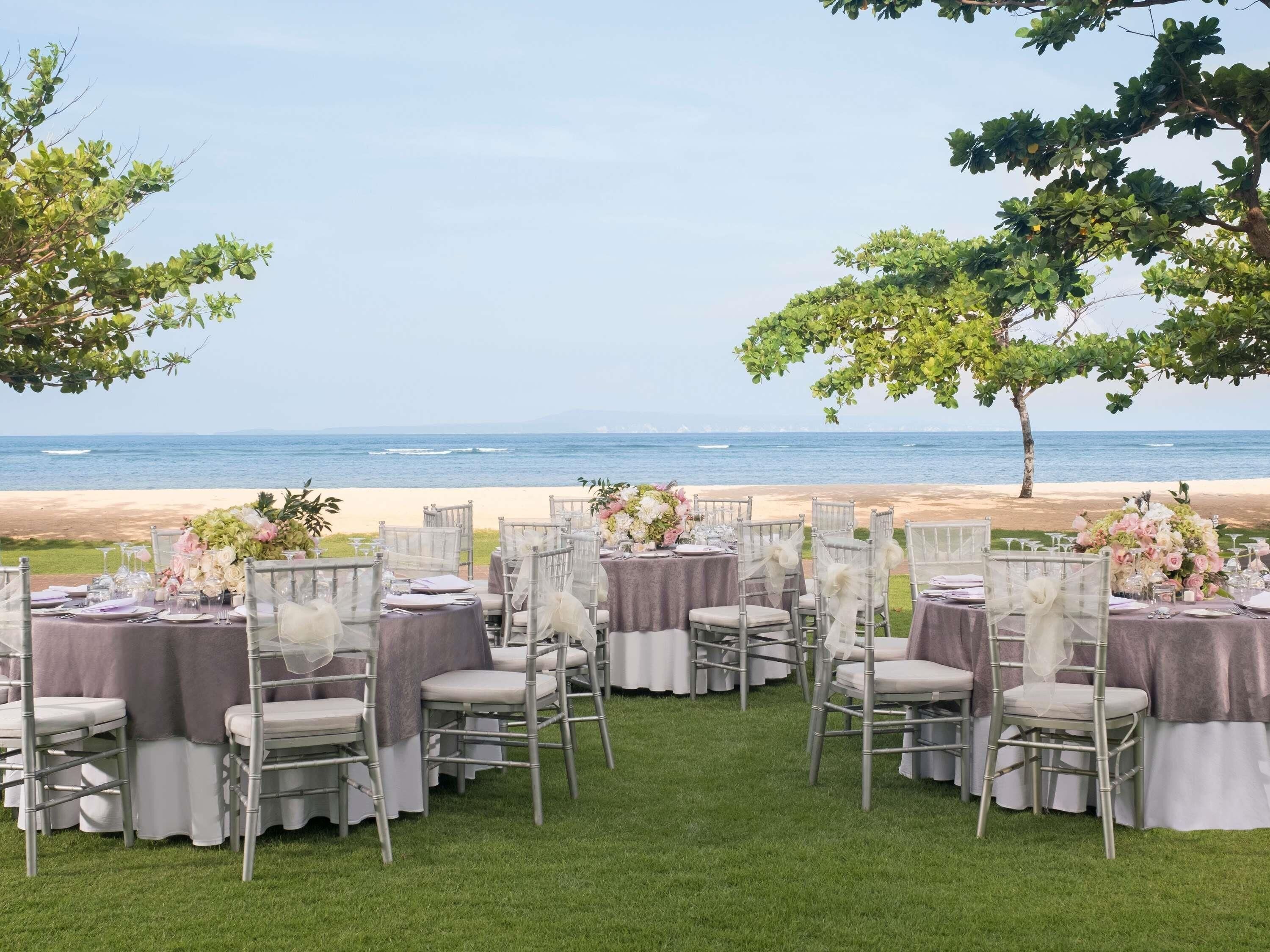 An outdoor wedding setup with tables and chairs arranged on grass, overlooking a beach and ocean.