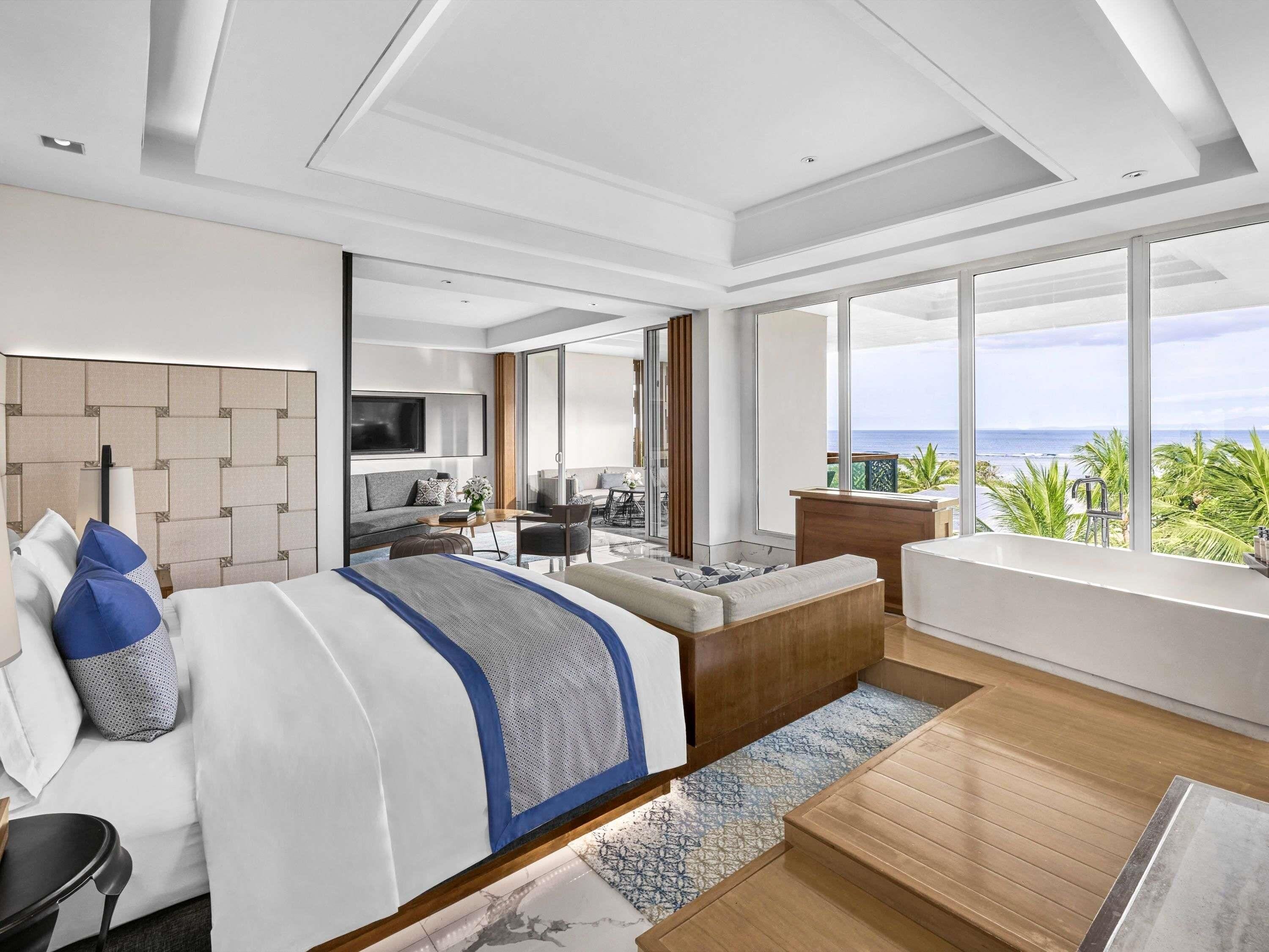 A modern hotel room with a large bed, a seating area, a balcony with a view of the ocean, and a TV mounted on the wall.