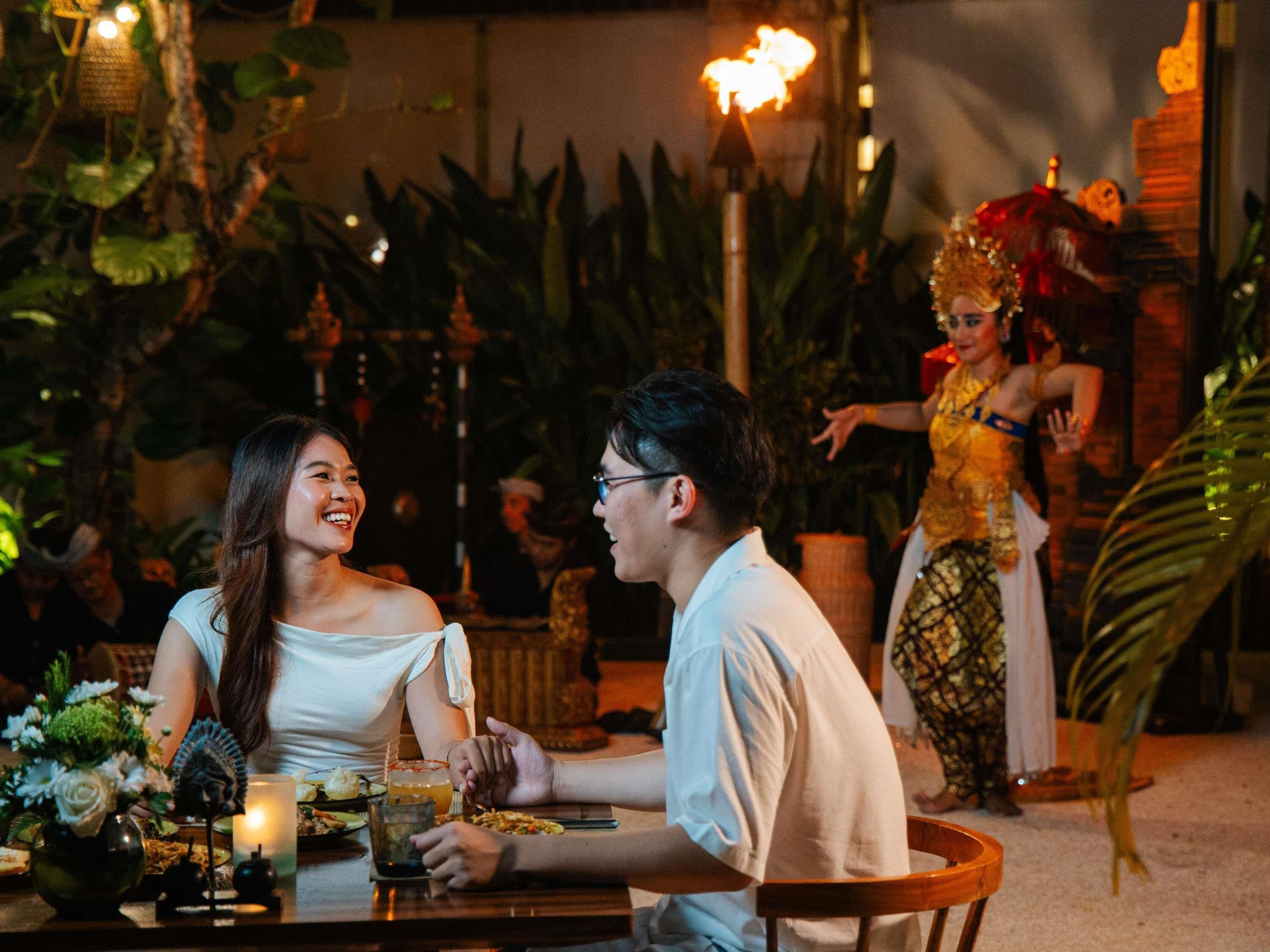 A dining area with a table set for two, surrounded by plants and candles, and a performer in traditional attire in the background.