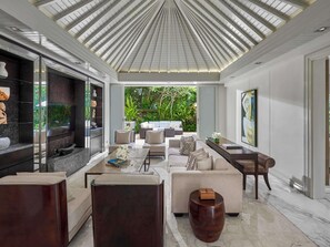 A modern living room with a large TV, comfortable seating, a coffee table, and a view of lush greenery outside.