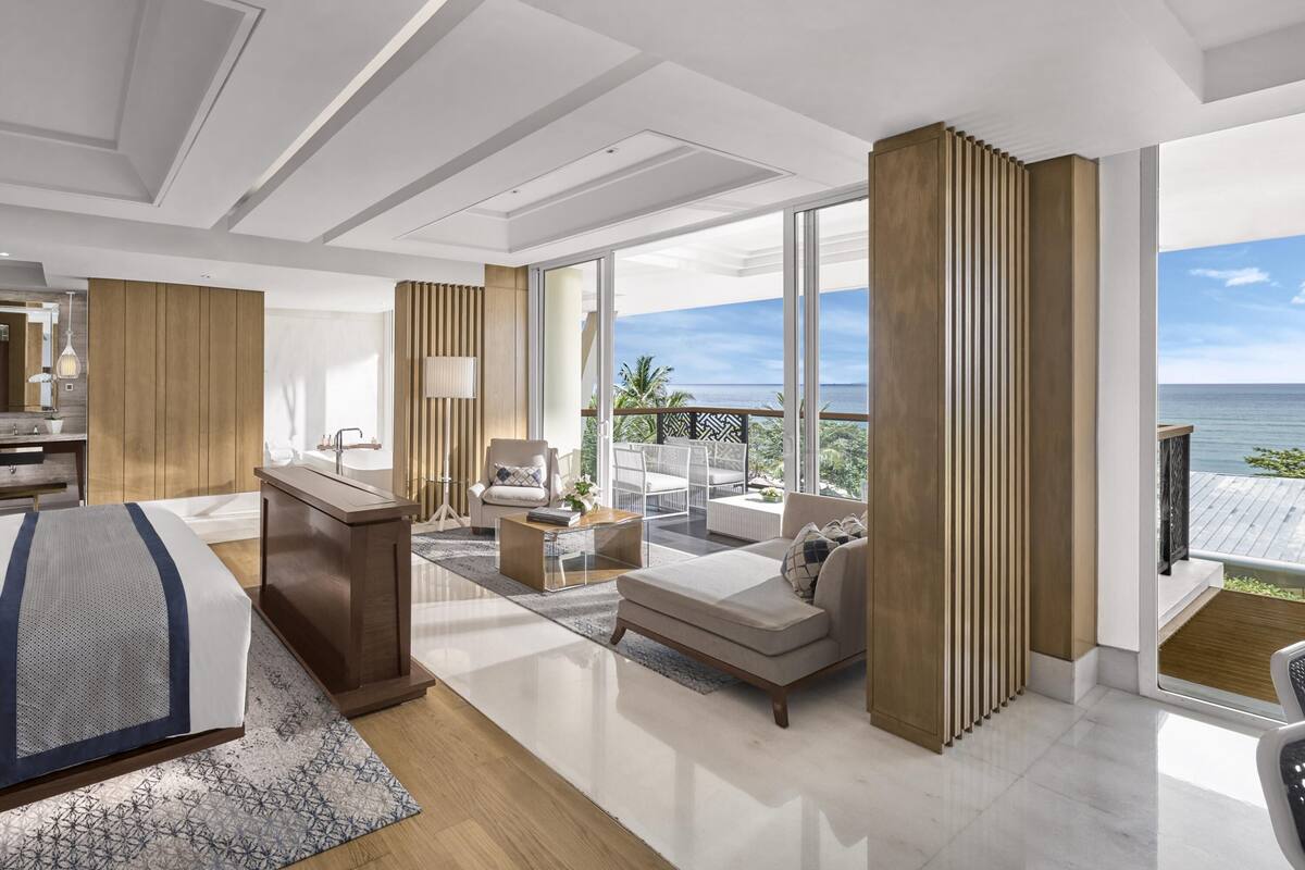 A modern hotel room with a large bed, a desk, a sofa, and a balcony with a view of the sea.