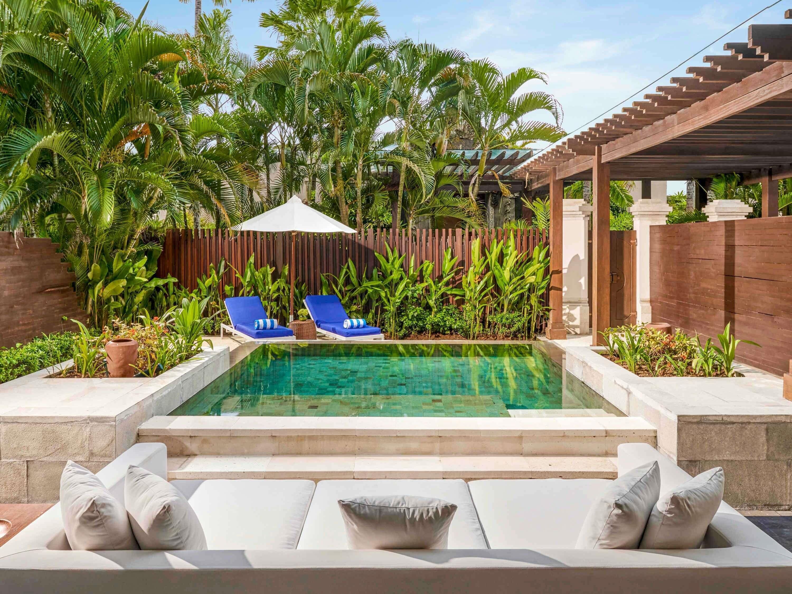 A swimming pool with lounge chairs and a covered area, surrounded by tropical plants and a wooden fence.