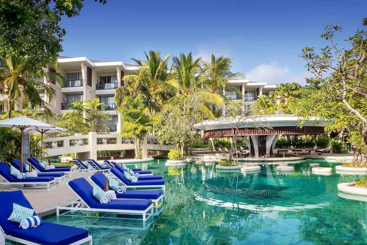 A resort pool with lounge chairs, palm trees, and a covered seating area.