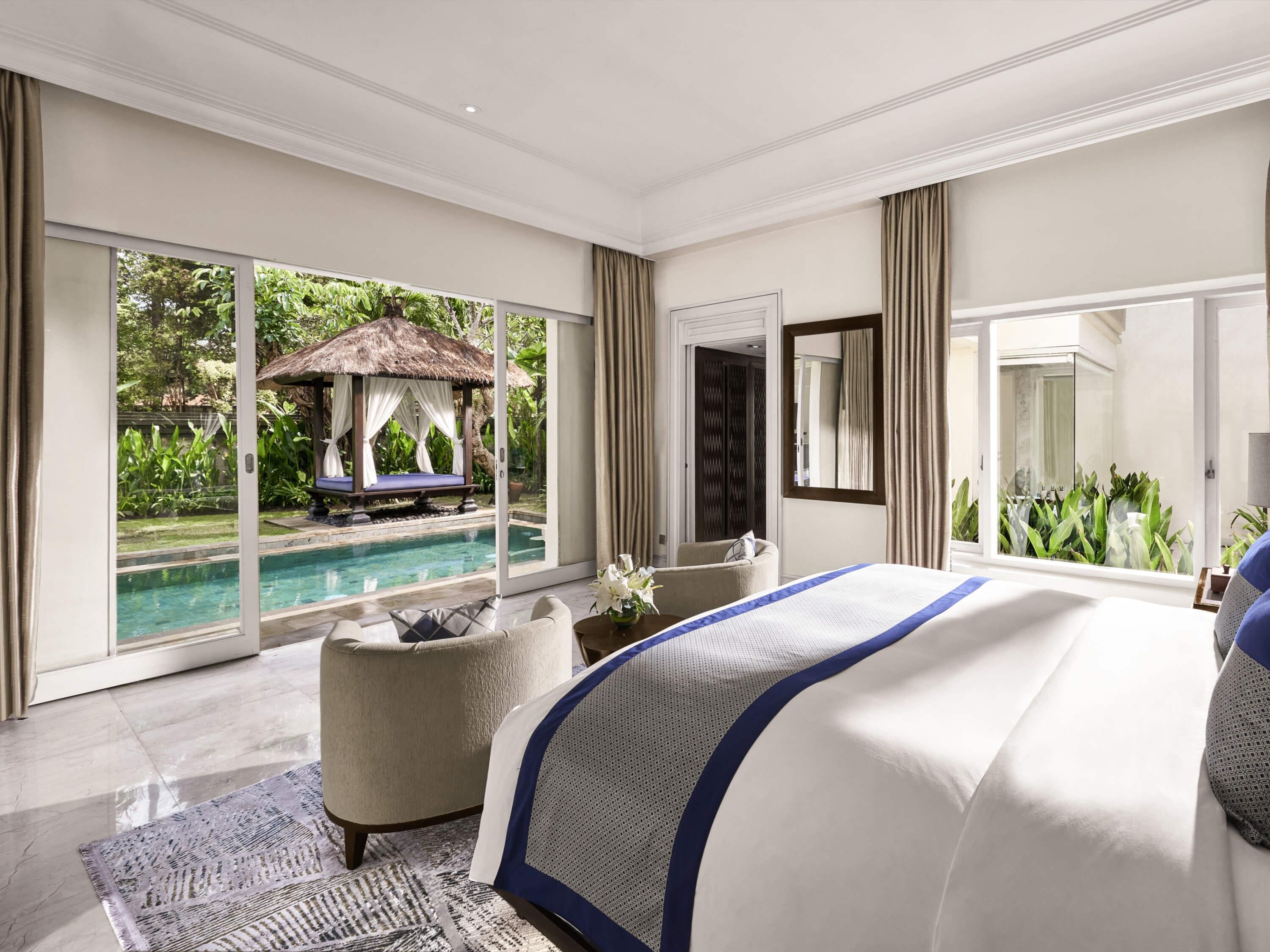 Ein Schlafzimmer mit einem großen Bett, einer Sitzecke, Blick auf einen Infinity-Pool und einem Reetdachgebäude.