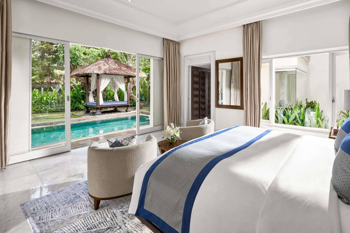 A spacious bedroom with a large bed, a view of a swimming pool, and a thatched-roof structure outside.