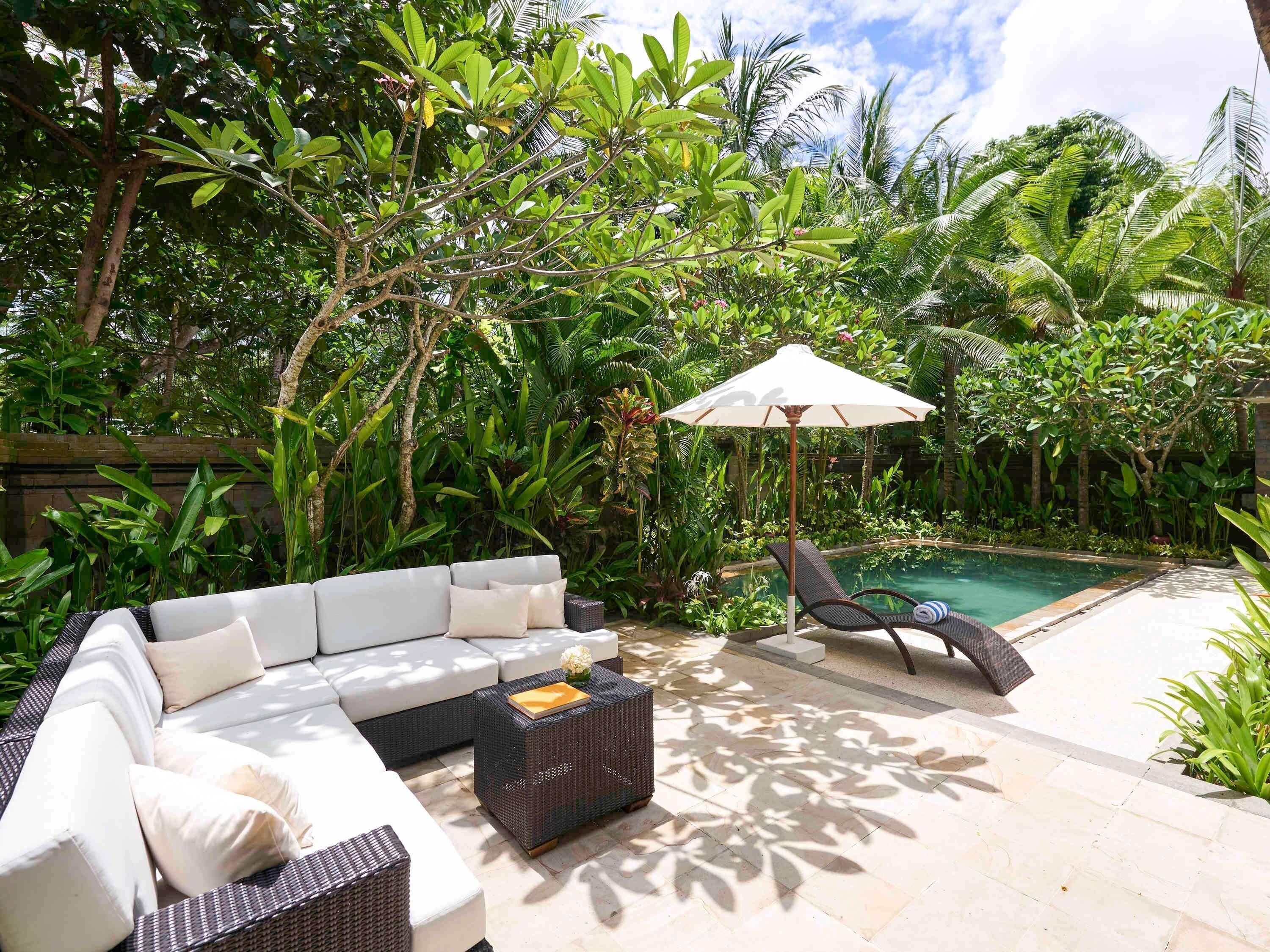 Un patio con un sofá blanco, una mesa de centro de mimbre, una piscina y abundante vegetación.
