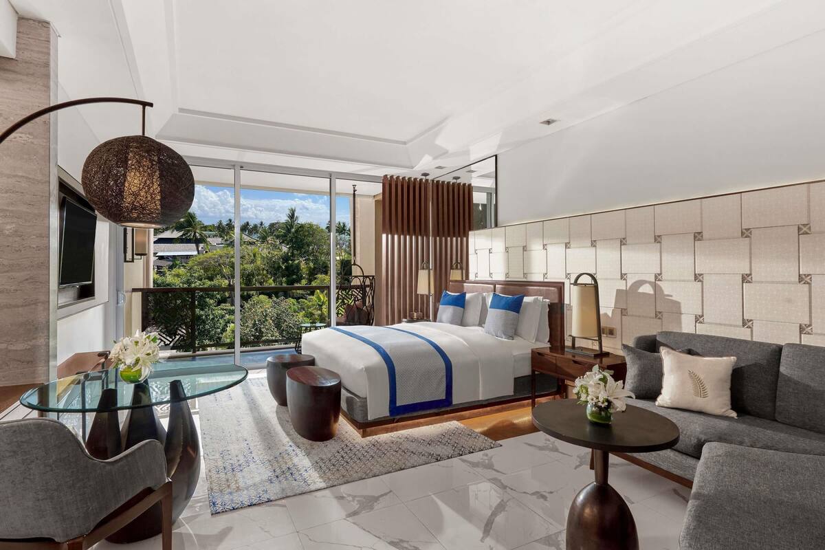 A modern hotel room with a large bed, a sofa, a glass table, and a balcony with a view.