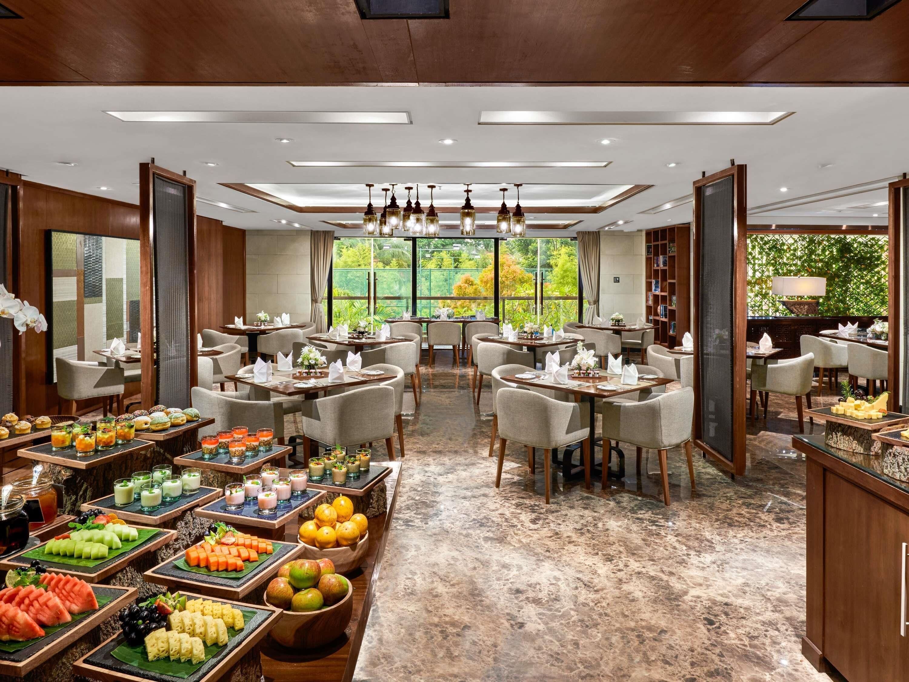 Un restaurante con varias mesas, un bufé de frutas y postres, y vistas al exterior con zonas verdes.