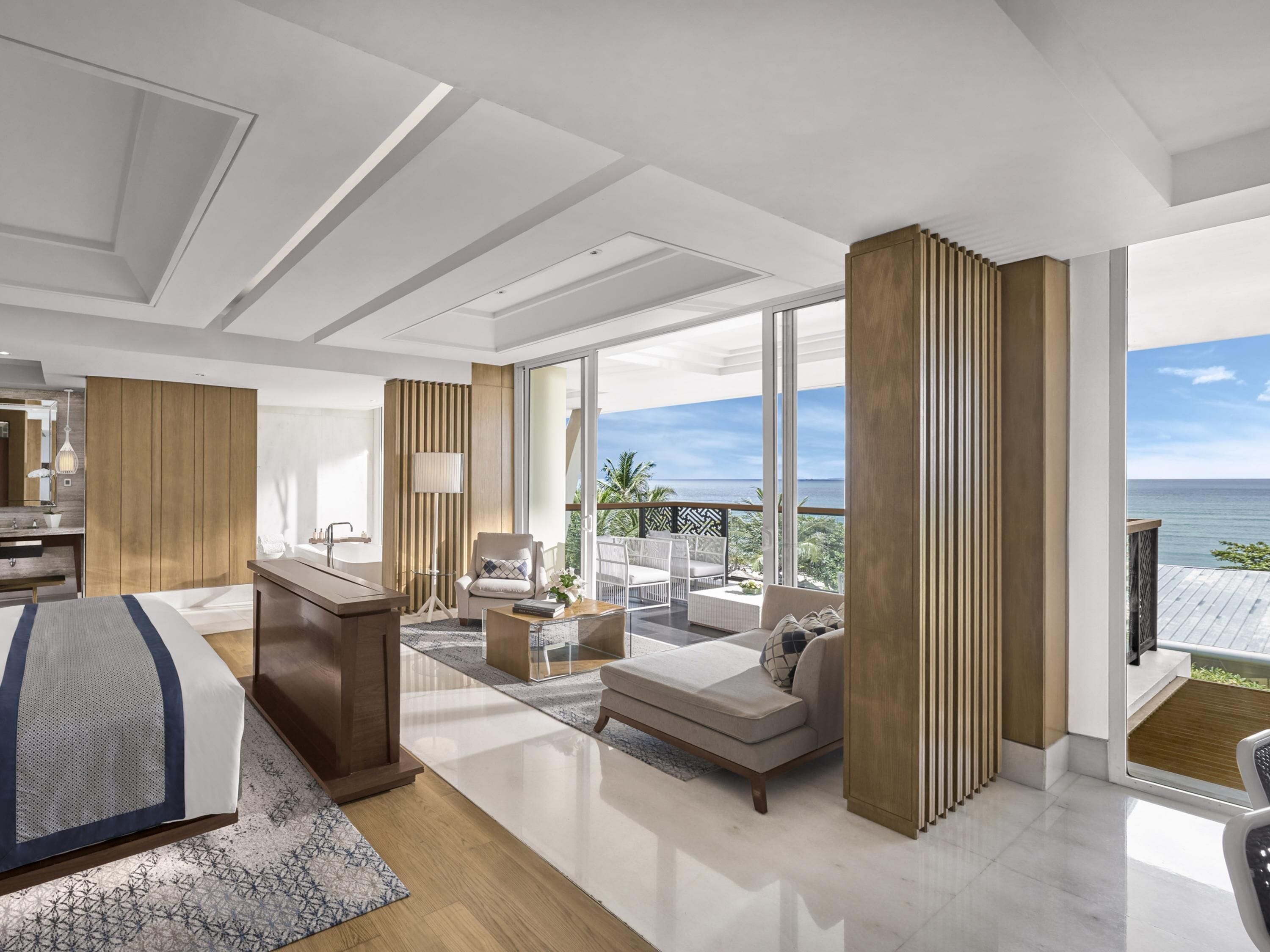 A modern hotel room with a large bed, a desk, a sofa, and a balcony with a view of the sea.