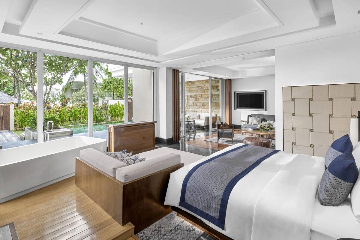 A modern bedroom with a large bed, a seating area with a view of the outdoors, and a TV mounted on the wall.