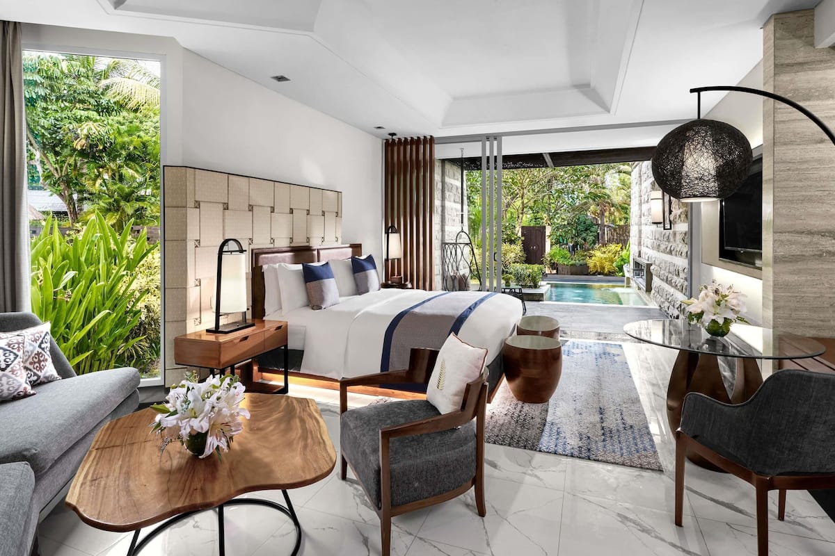 Une chambre d’hôtel moderne dotée d’un grand lit, d’un coin salon avec une table basse et des fauteuils, d’une télévision, et offrant une vue sur une piscine et un jardin.