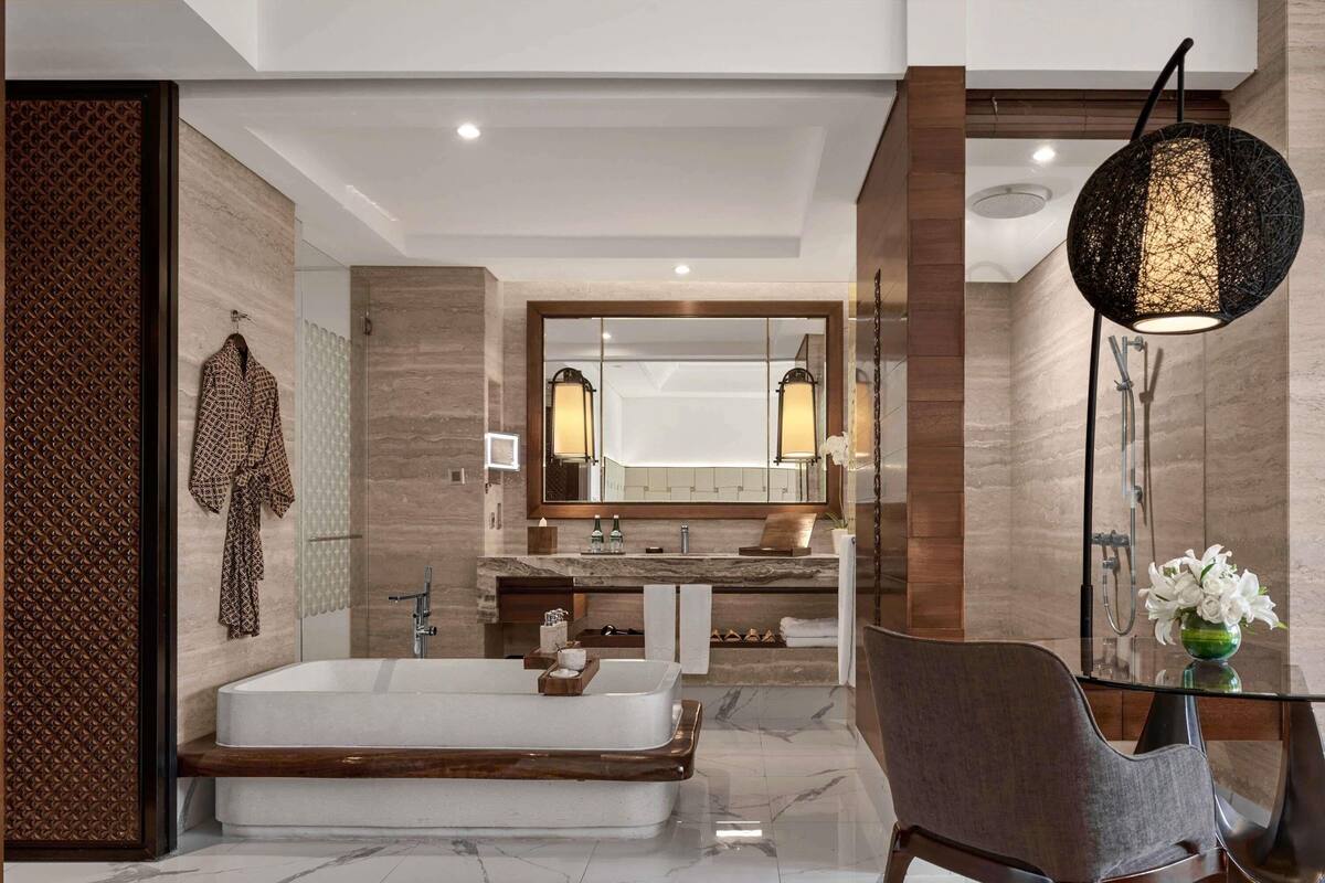 A modern bathroom with a large mirror, a bathtub, a vanity with a marble countertop, and a chair.