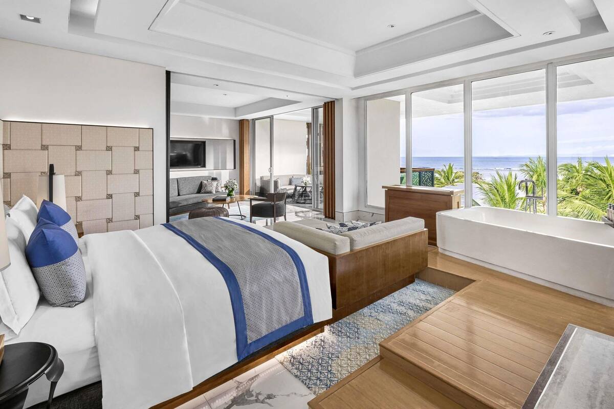 A modern hotel room with a large bed, a seating area, a balcony with a view of the ocean, and a TV mounted on the wall.