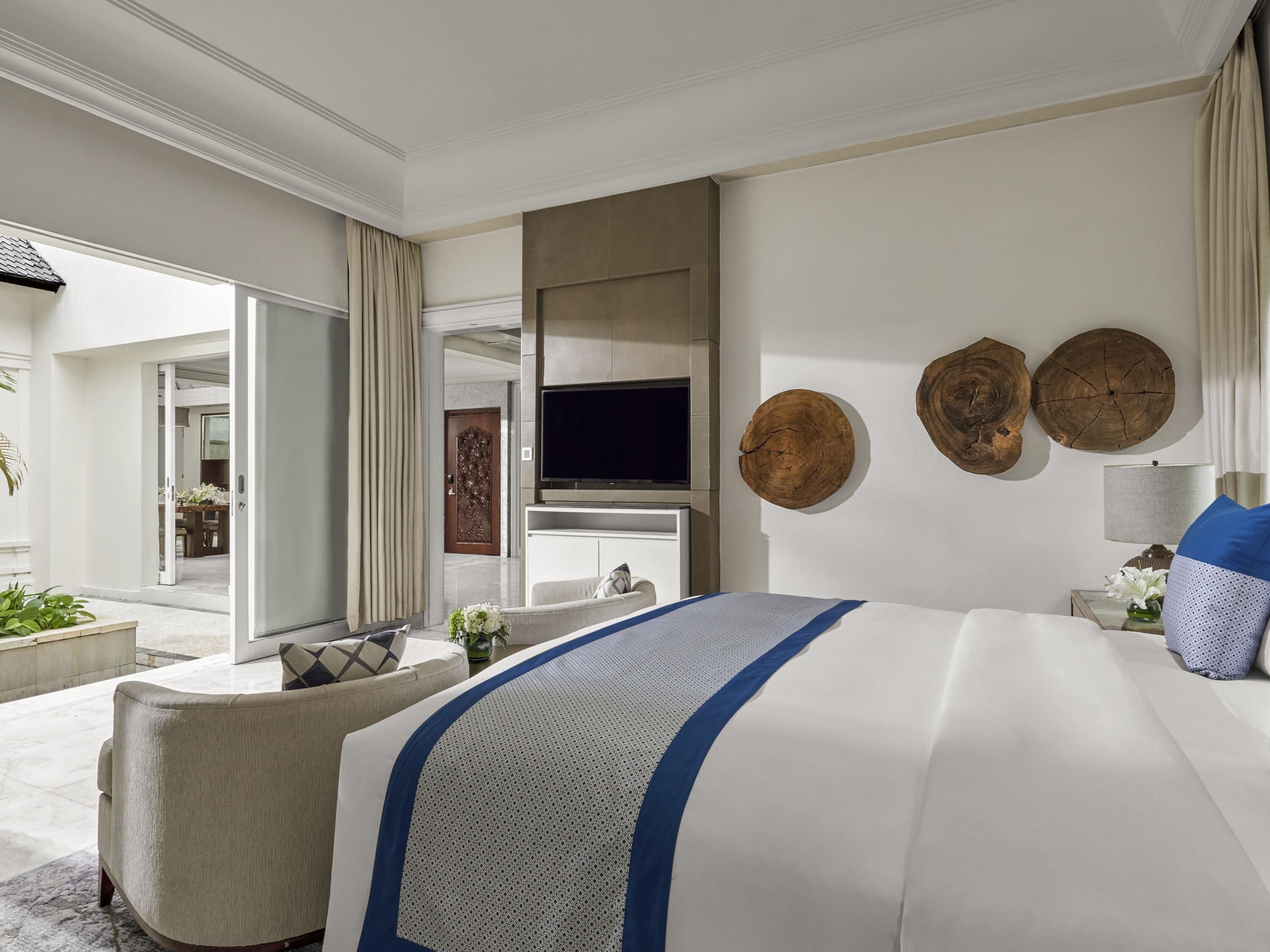 A modern hotel room with a large bed, a sofa, a TV, and a dining area visible through an open door.