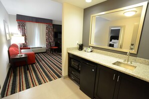 A compact hotel room with a kitchenette, a bed, a seating area, and a large mirror. - Hampton Inn & Suites Albany At Albany Mall (Albany)