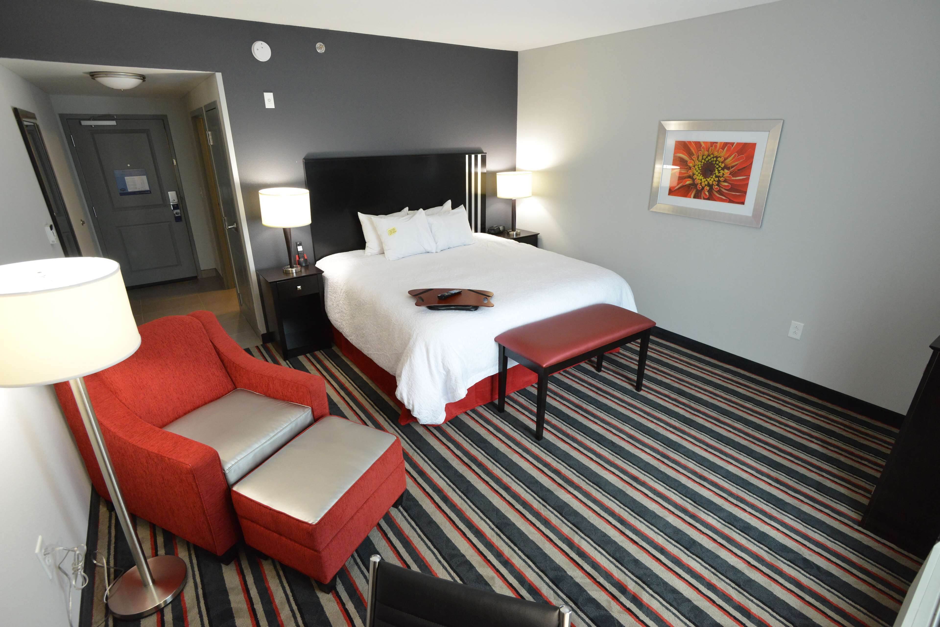 A hotel room with a bed, a red armchair, a bench, a nightstand with a lamp, and a framed picture on the wall.