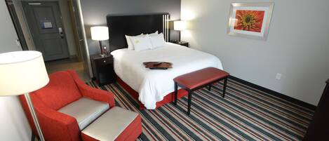 A hotel room with a bed, a red armchair, a bench, a nightstand with a lamp, and a framed picture on the wall.