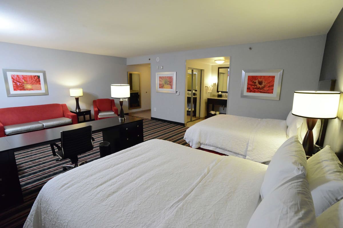 A hotel room with two beds, a red sofa, a desk with a chair, and a bathroom visible through an open door.