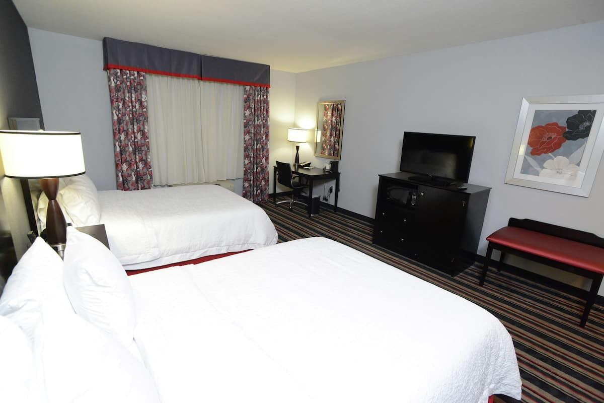 A hotel room with two beds, a desk, a television, a bench, and a painting on the wall.