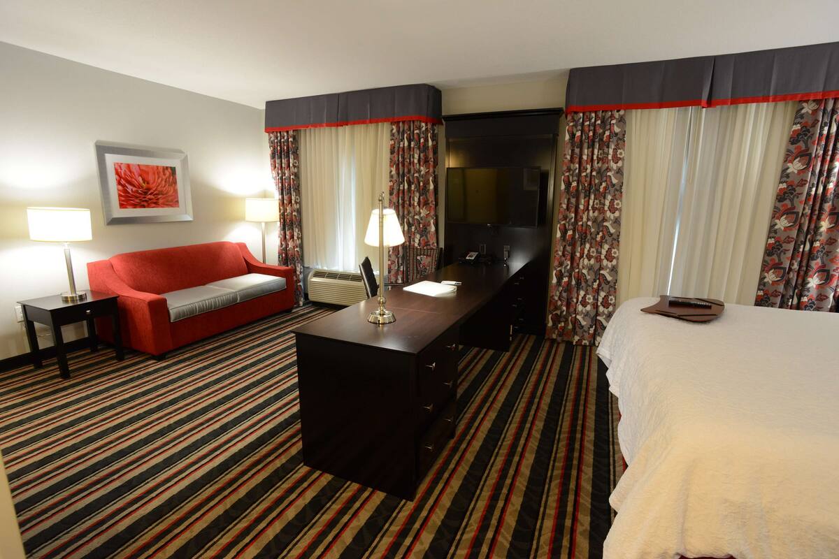 A hotel room with a red sofa, a desk with a lamp, a bed with a white cover, and floral curtains.
