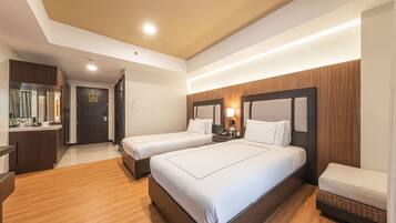 Habitación de hotel con dos camas, piso de madera, escritorio con espejo y baño visible a través de una puerta abierta.