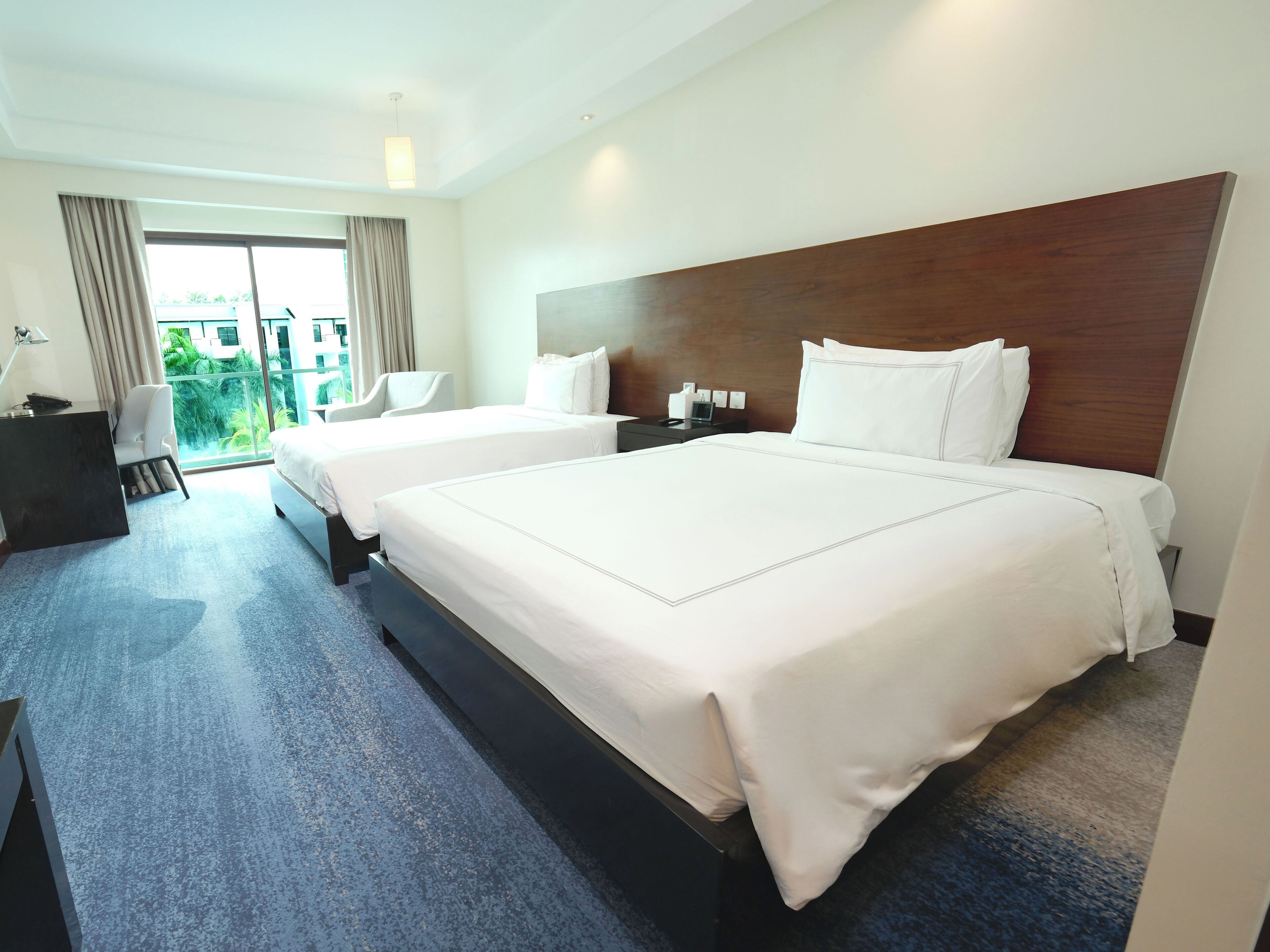 A hotel room with two beds, a desk, a chair, and a view of greenery outside.
