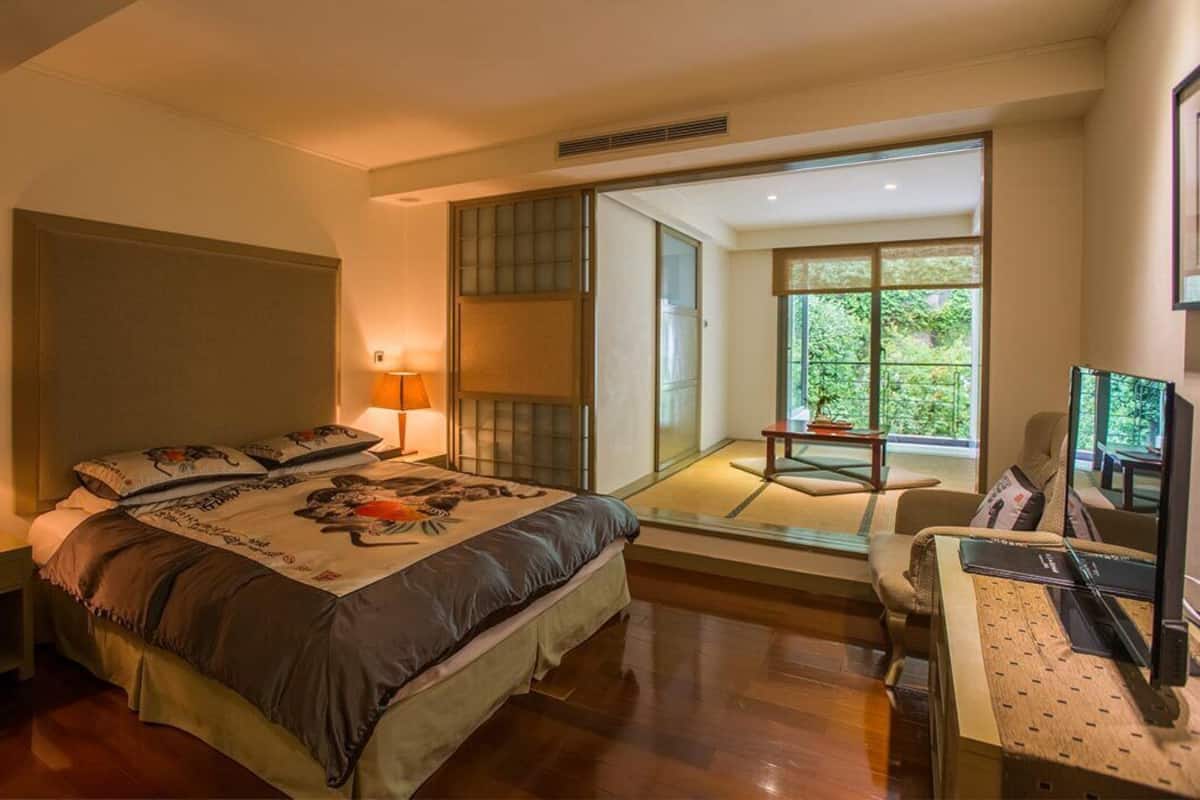 A bedroom with a bed, bedside table, lamp, and a view of a balcony with a chair and table.