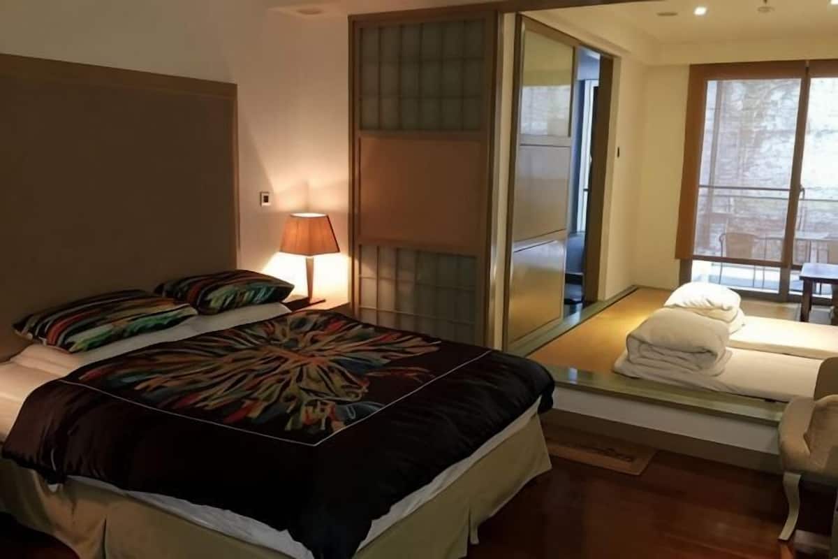 A bedroom with a bed, a nightstand, a lamp, a glass-topped table with towels, and a sliding door leading to a balcony.