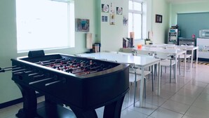 Una habitación con mesa de fútbol, mesas y sillas blancas, un reloj y una ventana.
