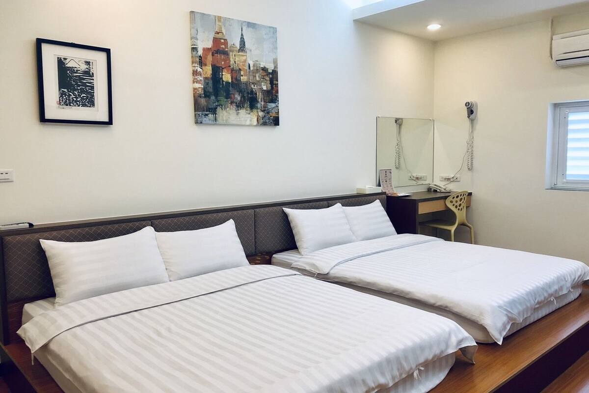 A hotel room with two single beds, a desk, a chair, a mirror, and two framed artworks on the wall.
