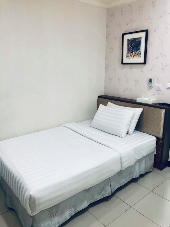 Un lit simple avec une couvre-lit blanche et des oreillers assortis, dans une pièce dont le papier peint est à motifs floraux et où un tableau encadré est accroché au mur.
