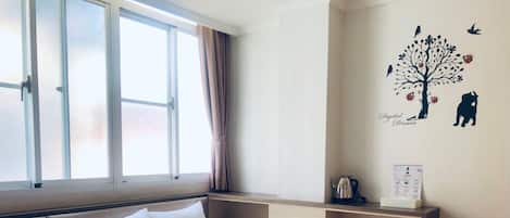 Habitación de hotel con cama, ventana con cortinas, decoración en la pared y una mesita con hervidor y cartel.