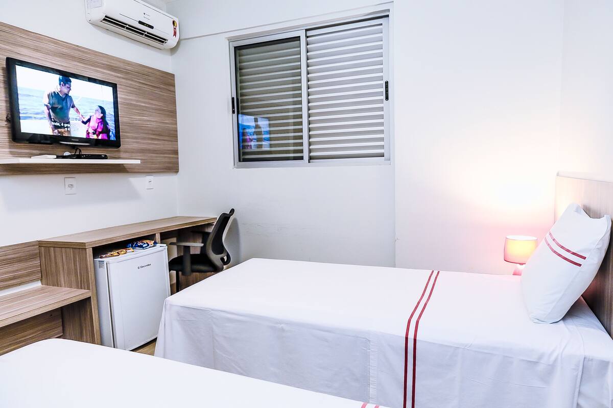 Une chambre d’hôtel comprenant un lit, un bureau avec un ordinateur, une télévision, une fenêtre avec des stores et un réfrigérateur.