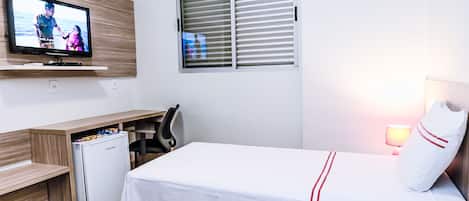 A hotel room with a bed, a desk with a computer, a television, a window with blinds, and a refrigerator.
