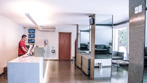 A modern reception area with a receptionist, a reception desk, a seating area with sofas, and a large mirror reflecting the space. - Hotel Rio Vermelho (Goiania)
