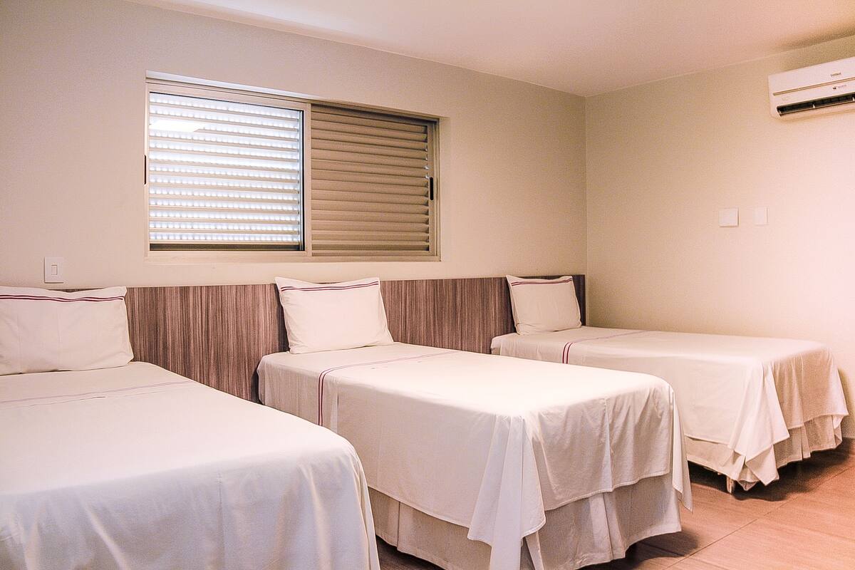 Une chambre d’hôtel avec trois lits simples, une fenêtre avec des stores et un appareil de climatisation.